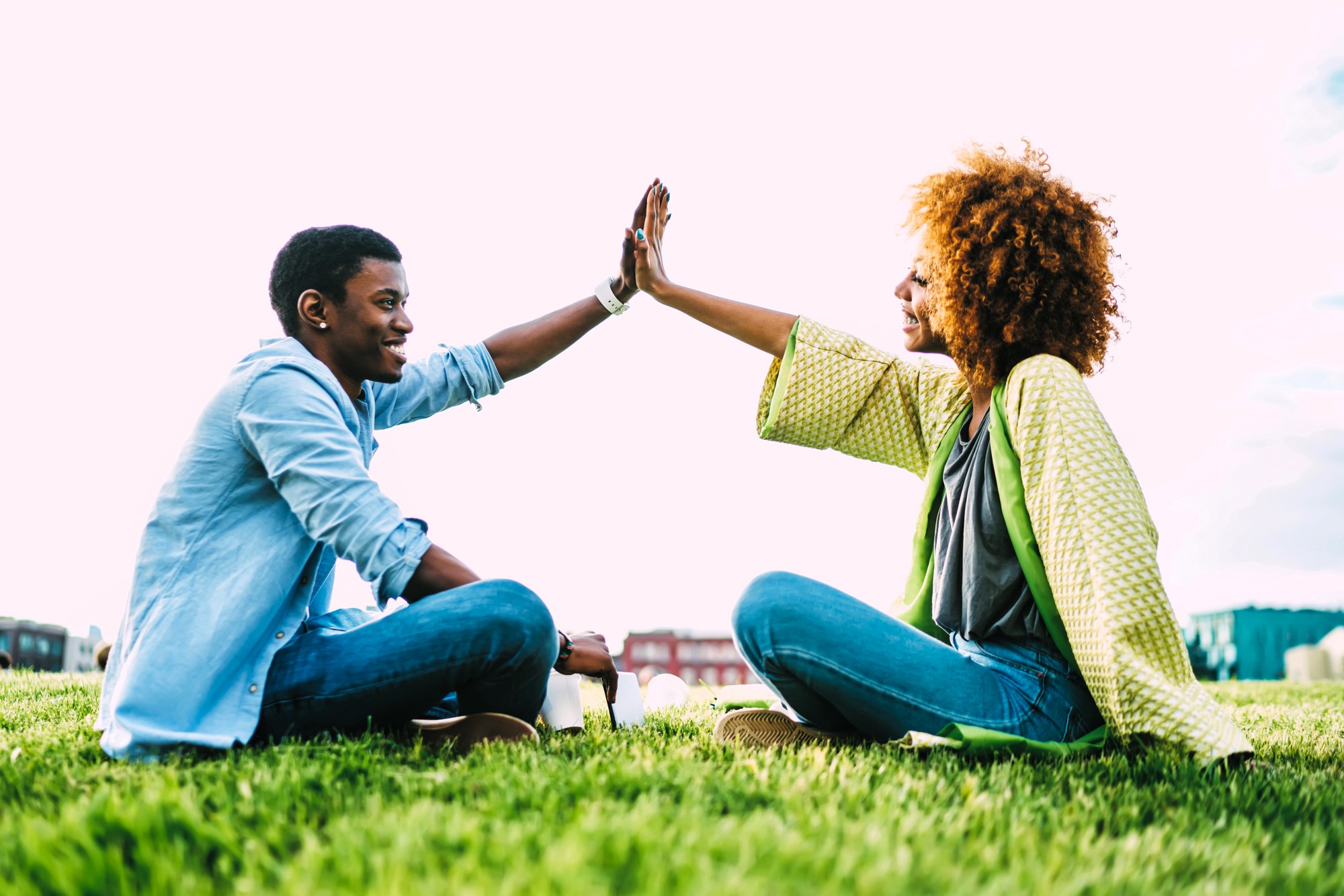 Two people sitting on grass, facing each other, giving a high-five. One wears a light blue shirt; the other wears a green patterned jacket. Both are smiling and appear to be enjoying a sunny day in an open area.