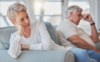 A woman with short gray hair sits on a couch, resting her head on her hand, looking away thoughtfully. In the background, an older man with white hair sits with his hand on his forehead, appearing pensive.