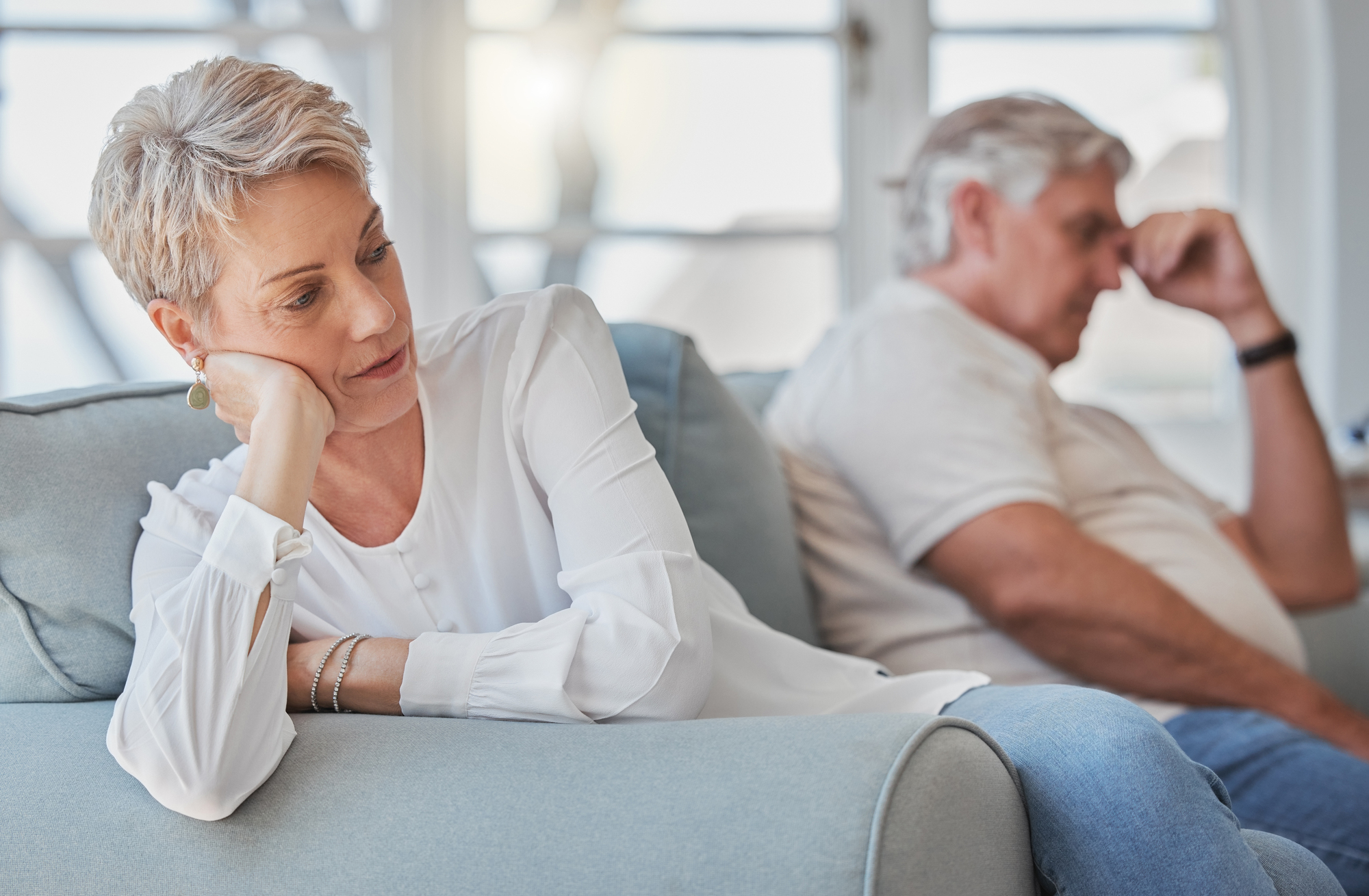 A woman with short gray hair sits on a couch, resting her head on her hand, looking away thoughtfully. In the background, an older man with white hair sits with his hand on his forehead, appearing pensive.