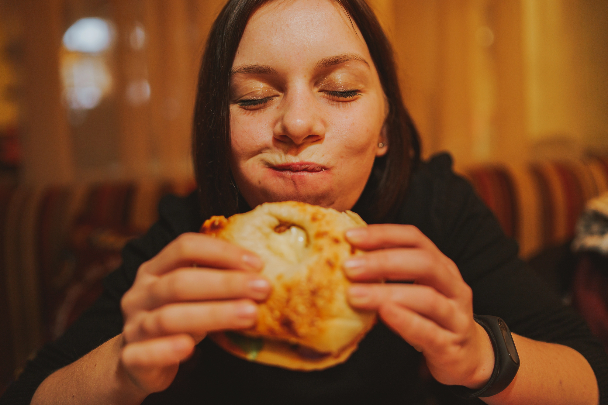 A person with brown hair is happily eating a sandwich indoors. They are wearing a black sweater and a watch, with their eyes closed and a content expression. The background features soft, warm lighting.