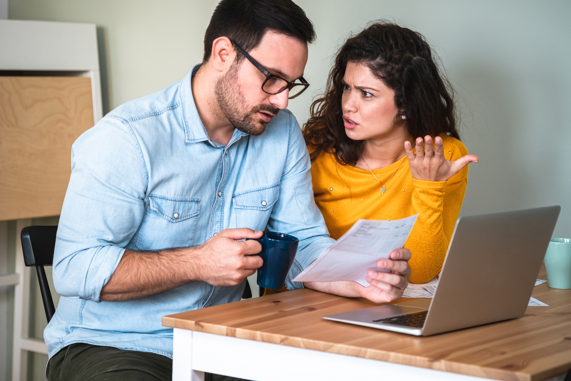 A man and woman sit at a table with a laptop, drinking from mugs. The man is reading a document, while the woman gestures in frustration, suggesting a discussion or disagreement about the paperwork.