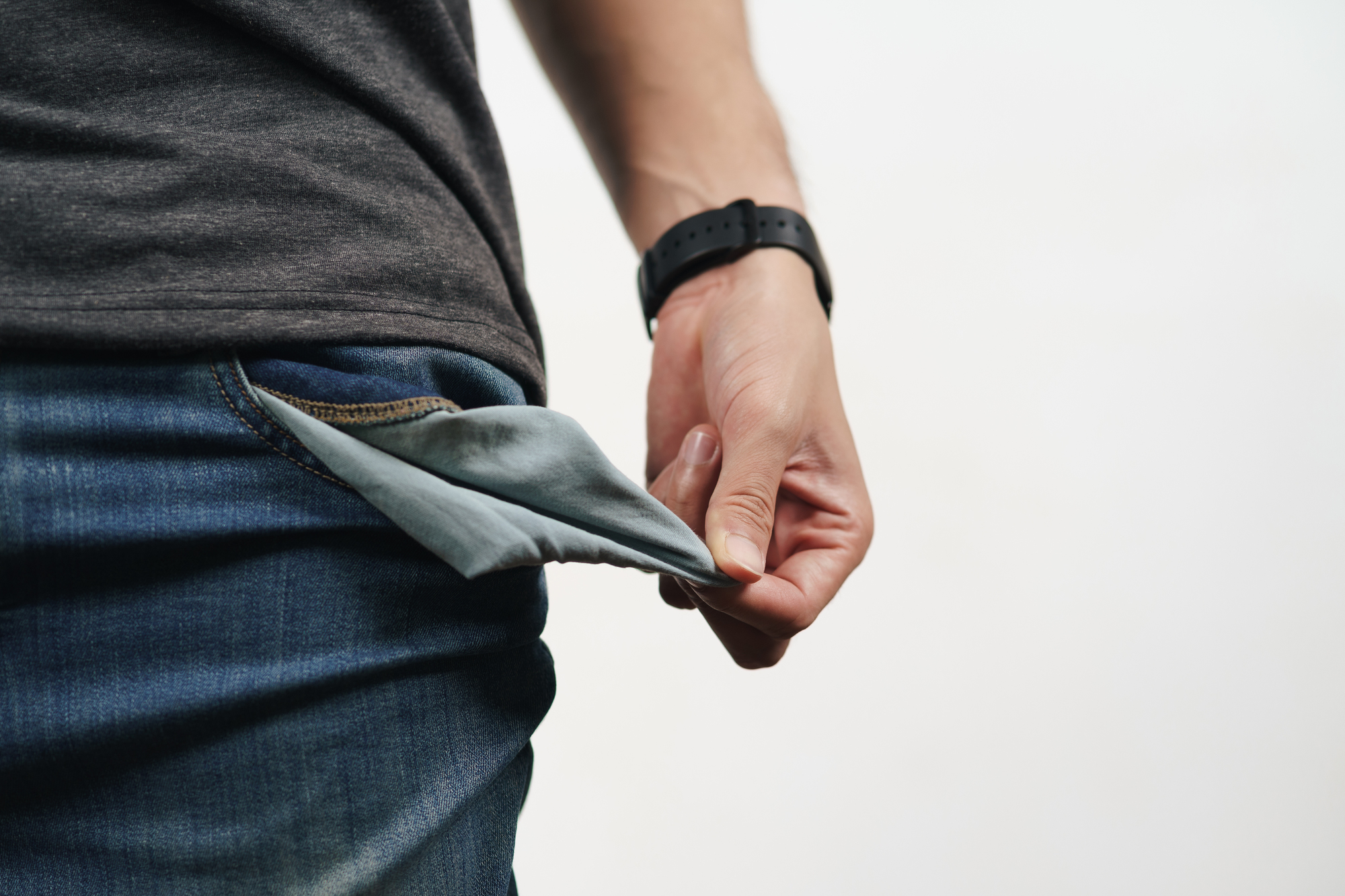 A person wearing a black wristwatch pulls out an empty pocket from their jeans, indicating no money. The focus is on the pocket and hand, with a plain white background.