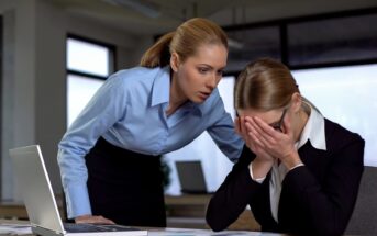 A woman in a blue shirt leans over, appearing concerned, as she looks at another woman in a black suit who is sitting at a desk with her head in her hands. An open laptop and documents are on the desk.