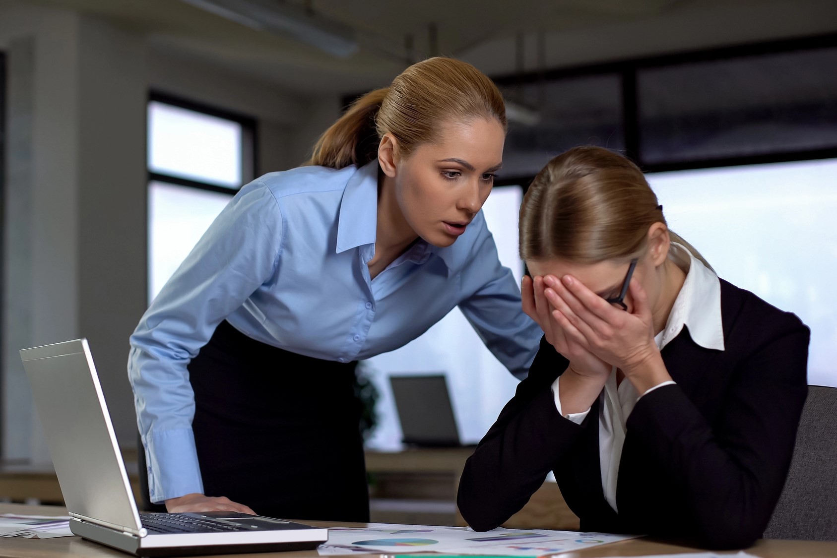 A woman in a blue shirt leans over, appearing concerned, as she looks at another woman in a black suit who is sitting at a desk with her head in her hands. An open laptop and documents are on the desk.