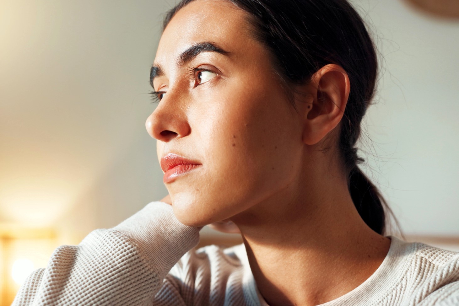 A person with long dark hair pulled back is looking contemplatively into the distance. They are indoors, wearing a light-colored sweater, with sunlight softly illuminating their face from the side.