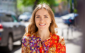 A smiling woman with long brown hair wearing a colorful floral blouse stands outdoors on a sunny day. The background shows a blurred street scene with cars and trees.