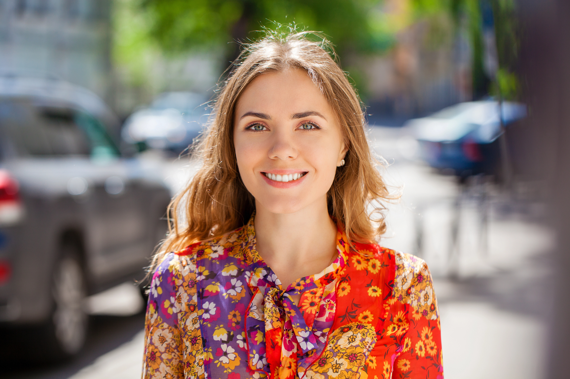 A smiling woman with long brown hair wearing a colorful floral blouse stands outdoors on a sunny day. The background shows a blurred street scene with cars and trees.