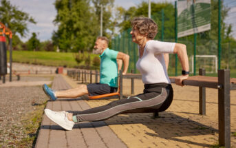 Two older adults exercising outdoors in a park on a sunny day. They are doing tricep dips using benches. Both are wearing athletic clothing, and the scene is set against a backdrop of greenery and a clear sky.