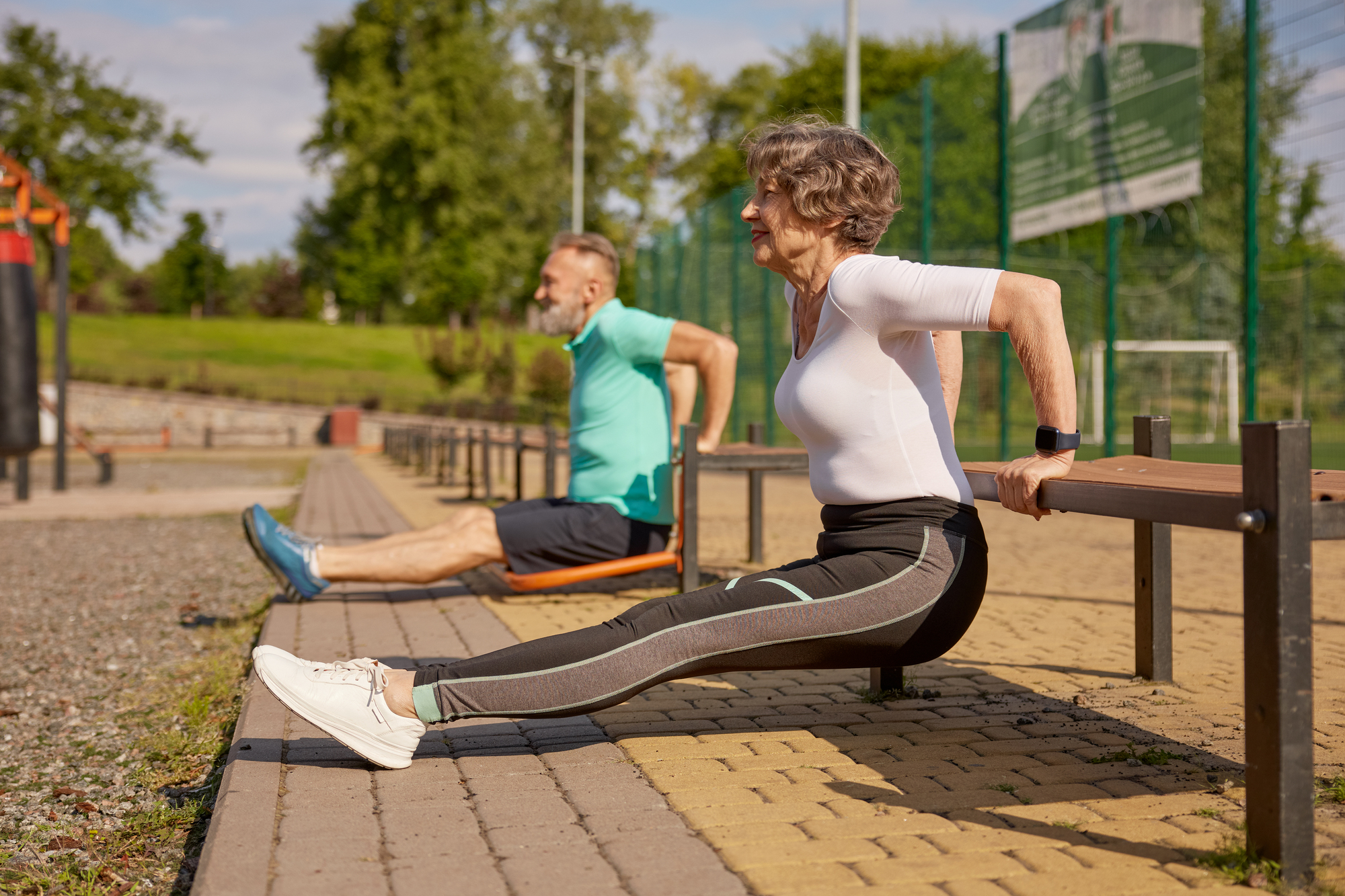 Two older adults exercising outdoors in a park on a sunny day. They are doing tricep dips using benches. Both are wearing athletic clothing, and the scene is set against a backdrop of greenery and a clear sky.