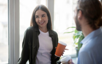 A woman in a white shirt and green jacket smiles while holding an orange coffee cup, standing by a window. She is engaged in conversation with a man wearing glasses holding a white cup. Bright, indoor setting with plants in the background.