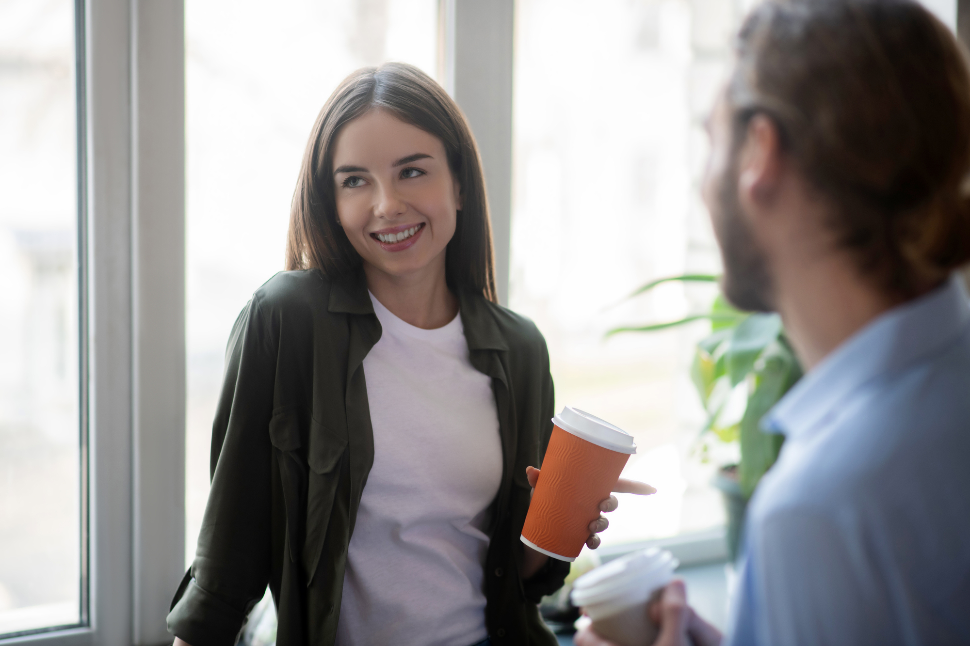 A woman in a white shirt and green jacket smiles while holding an orange coffee cup, standing by a window. She is engaged in conversation with a man wearing glasses holding a white cup. Bright, indoor setting with plants in the background.