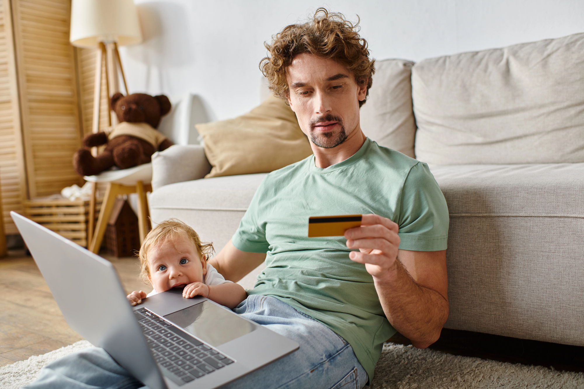 A man sits on the floor leaning against a couch, holding a credit card and using a laptop. A baby rests on his lap, looking at the computer screen. A teddy bear, lamp, and cushions are in the background.