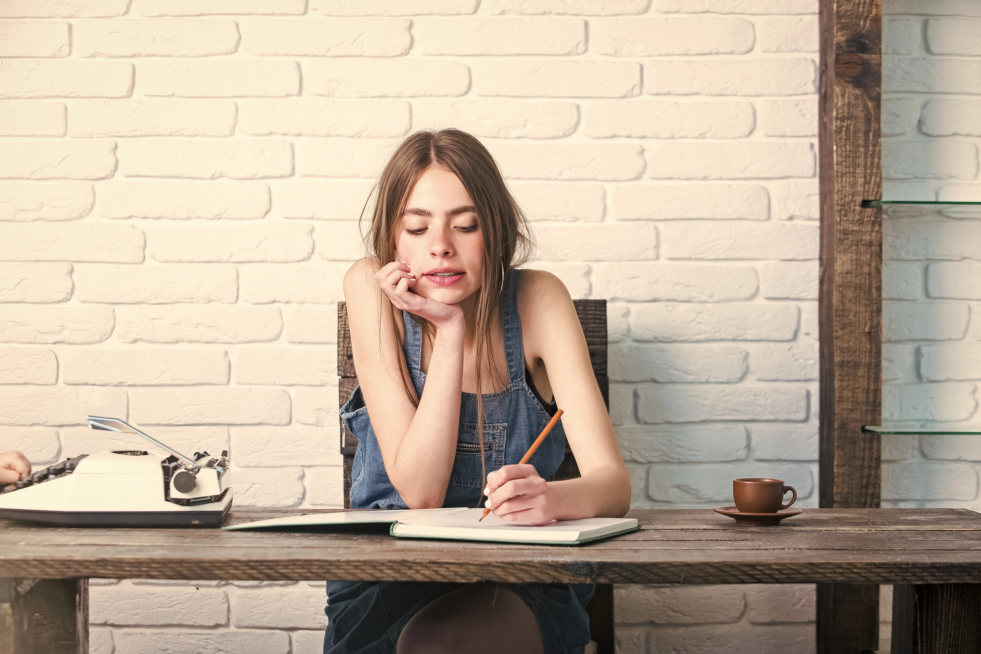 Woman in denim overalls sits at a wooden desk, thoughtfully writing in a notebook. A vintage typewriter and a cup of coffee are also on the desk. The background features a white brick wall.