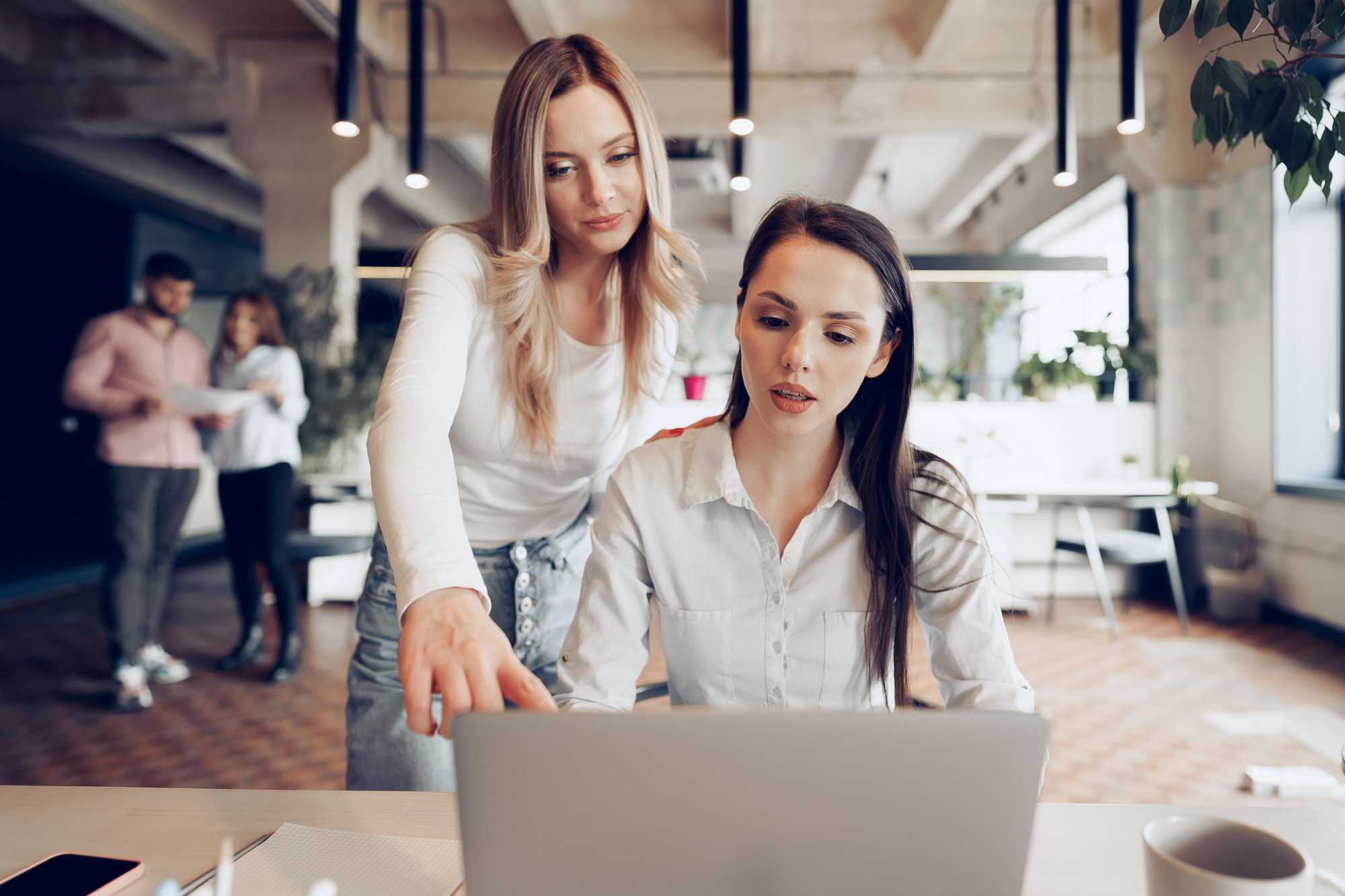 Two women at a desk in an open office setting. One is standing, pointing at a laptop screen, while the other is seated, looking at the screen. In the background, two people are having a conversation.