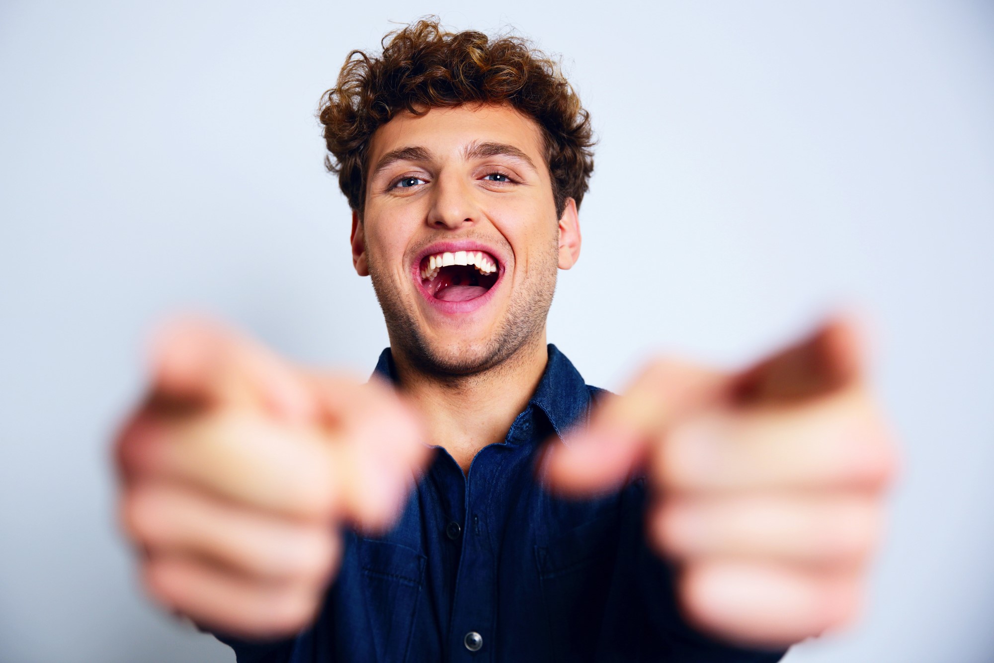 A young man with curly hair laughs energetically while pointing both index fingers towards the camera. He wears a dark blue shirt, and the background is a solid light color.