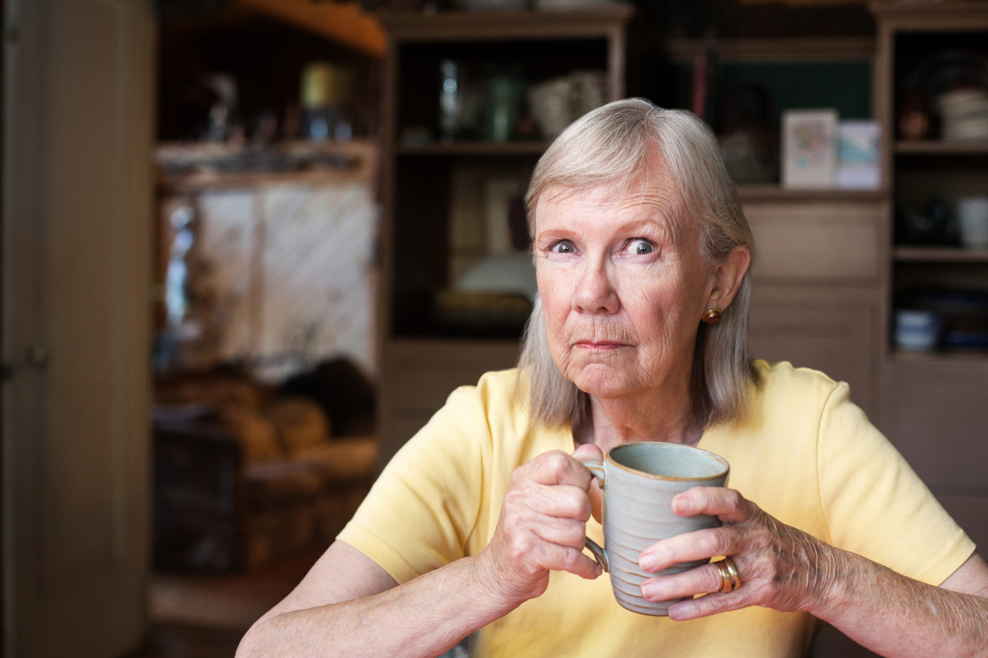 An elderly woman with gray hair and in a yellow shirt holds a mug with both hands while sitting indoors. The background shows shelves and a cozy room setting. Her expression is thoughtful.