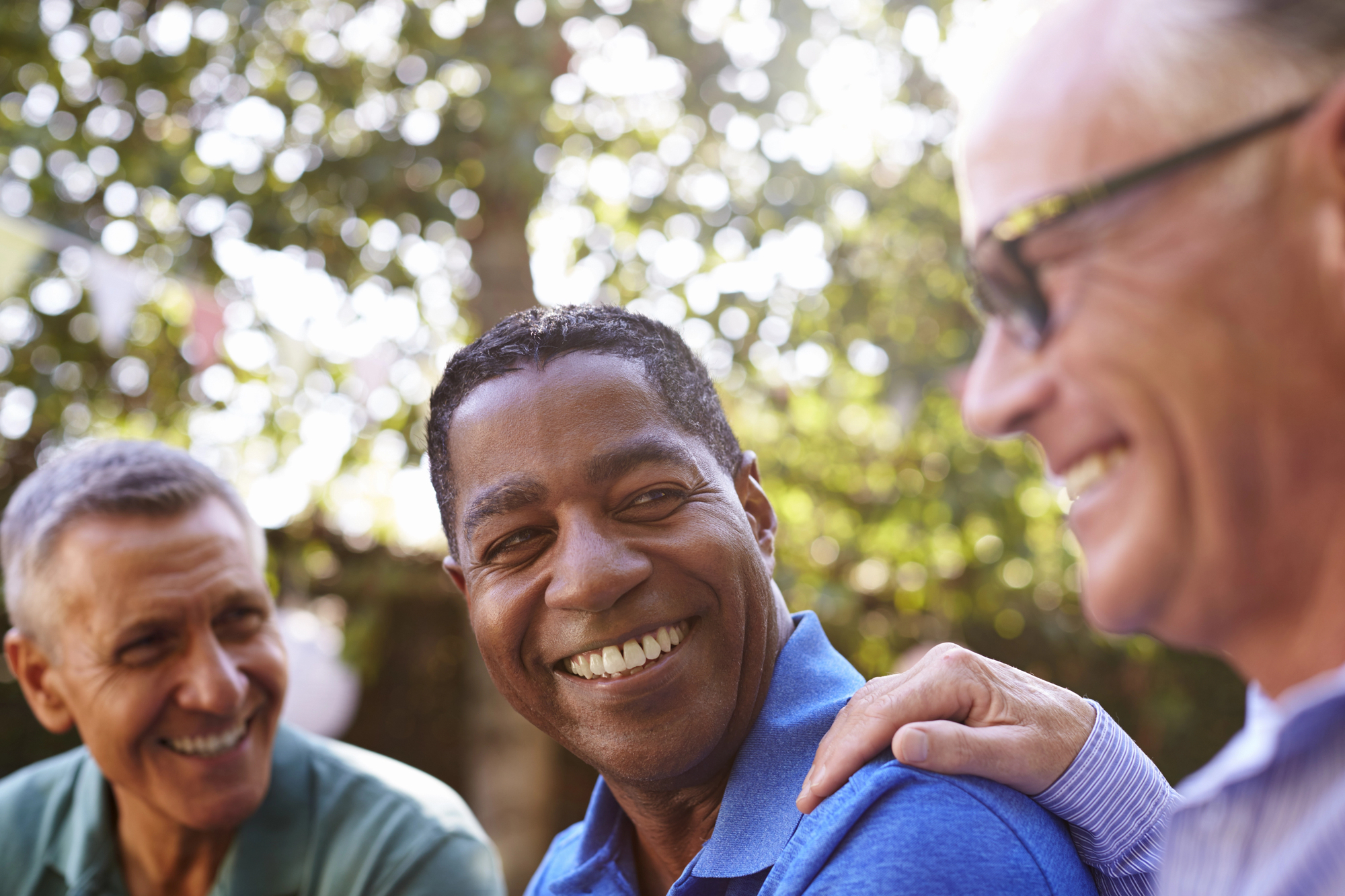 Three men are sitting outdoors and smiling warmly. The man in the middle is wearing a blue shirt, with the others on either side engaging with him. The background is a sunny, blurred garden setting with dappled light filtering through the trees.