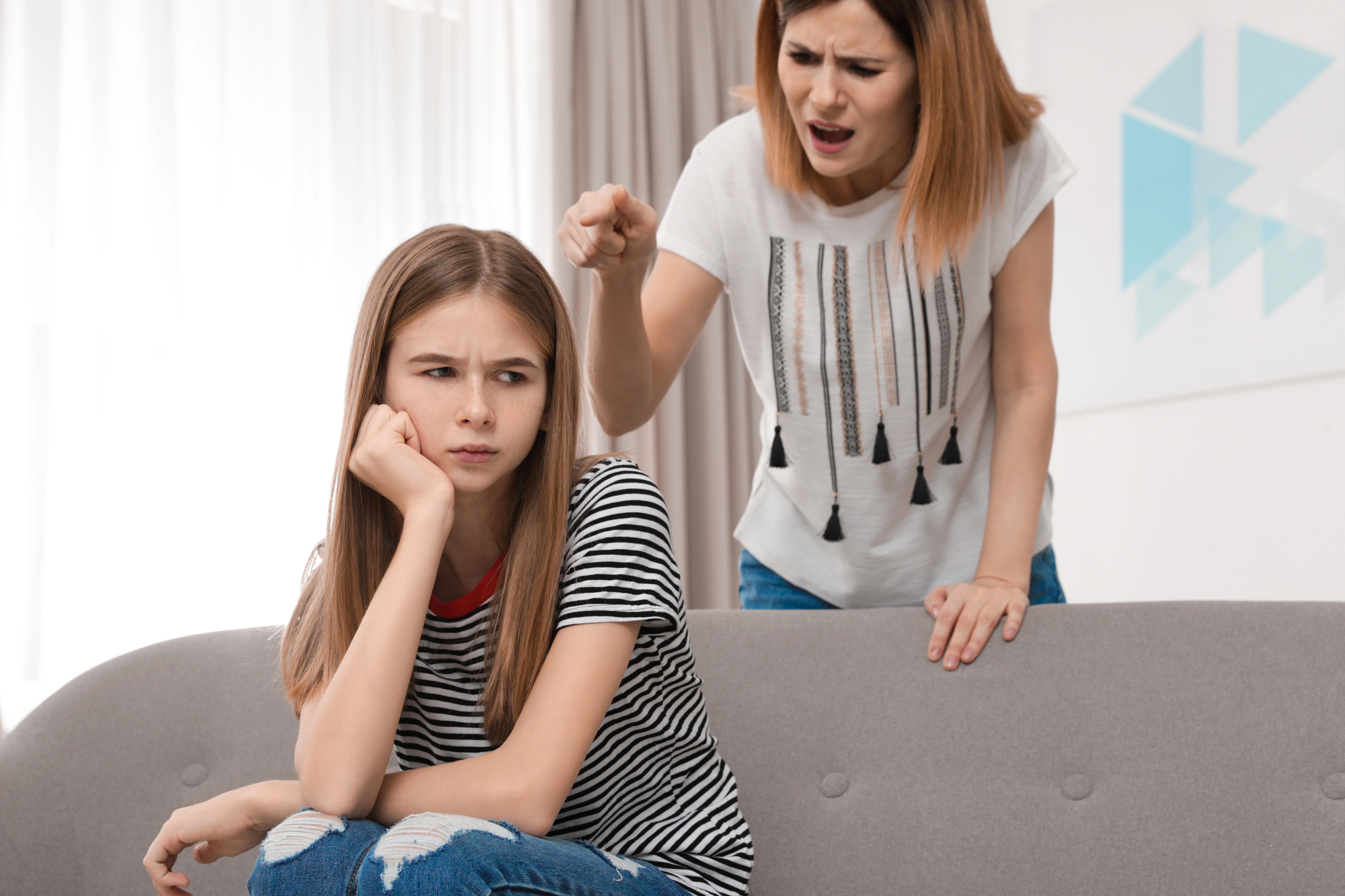 A woman appears to be scolding a girl, who looks away with a pensive expression. They are indoors, with the girl sitting on a couch and the woman standing behind her, pointing her finger. The atmosphere seems tense.