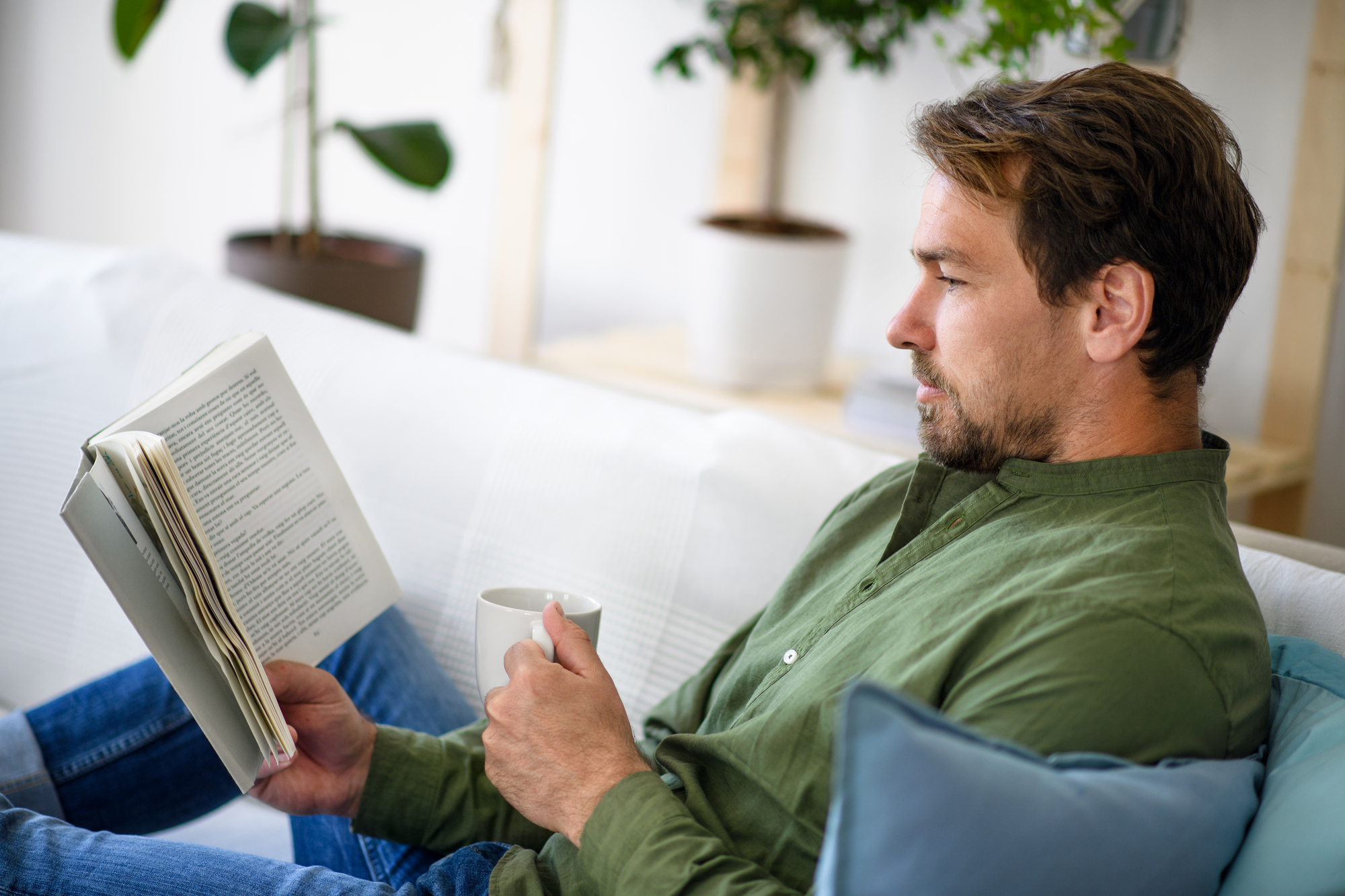 A man in a green shirt sits comfortably on a white couch, holding a mug in one hand and reading a book with the other. There are potted plants in the blurred background, adding a touch of greenery to the scene.