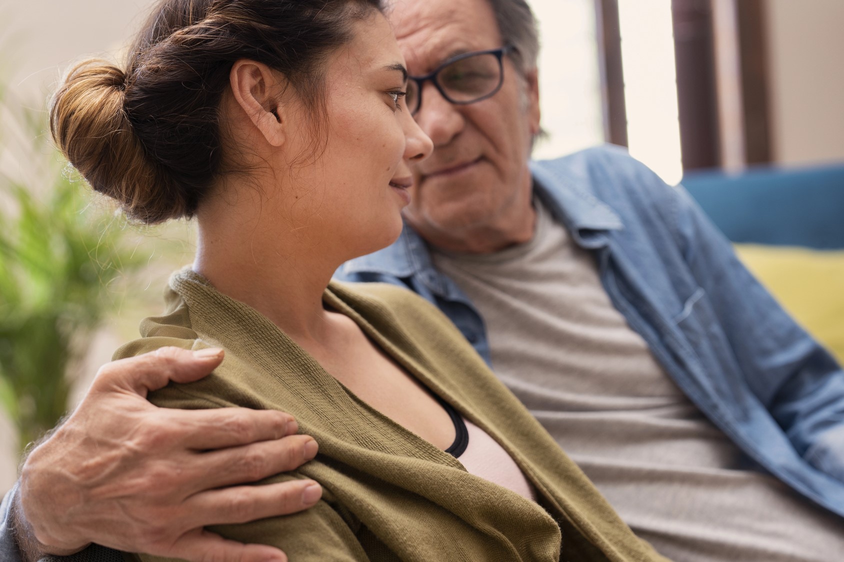 An older man with glasses sits closely beside a younger woman, gently embracing her with one arm. They are looking at each other, smiling warmly, in a cozy setting with soft lighting and a plant in the background.