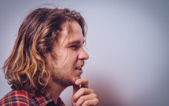 A young man with long, wavy hair and a beard is looking thoughtfully to the side. He is wearing a plaid shirt and has his hand to his chin in a pensive pose against a plain background.