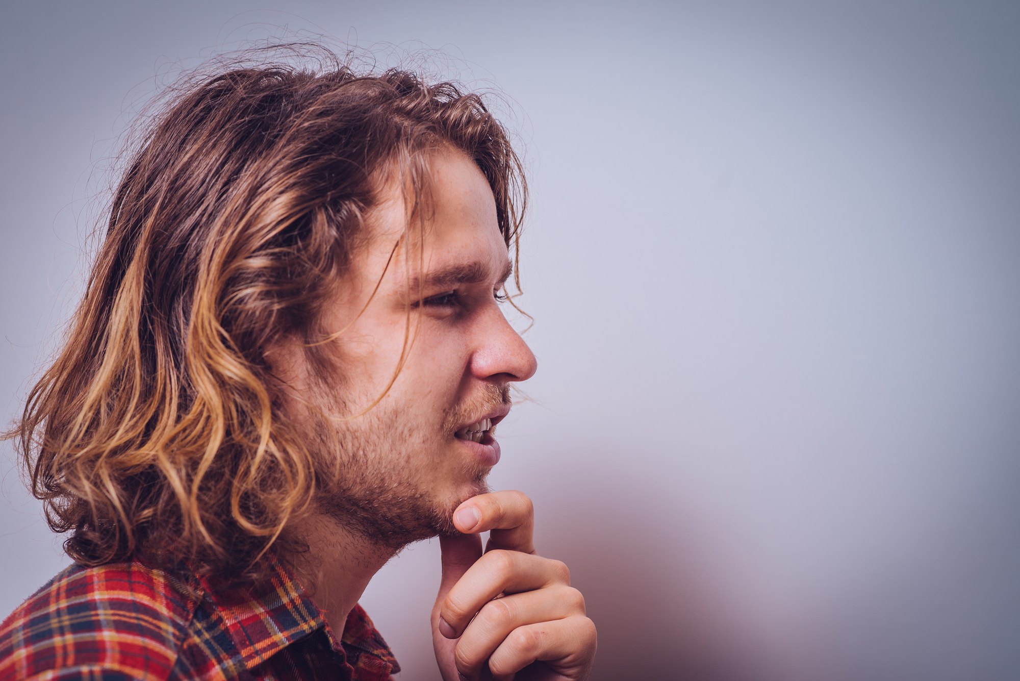 A young man with long, wavy hair and a beard is looking thoughtfully to the side. He is wearing a plaid shirt and has his hand to his chin in a pensive pose against a plain background.