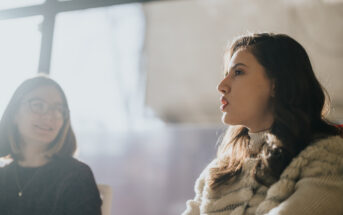 Two women are sitting and talking in a softly lit room. They are engaged in conversation, with one woman in focus as she speaks, while the other listens attentively. Natural light filters in from a large window behind them.