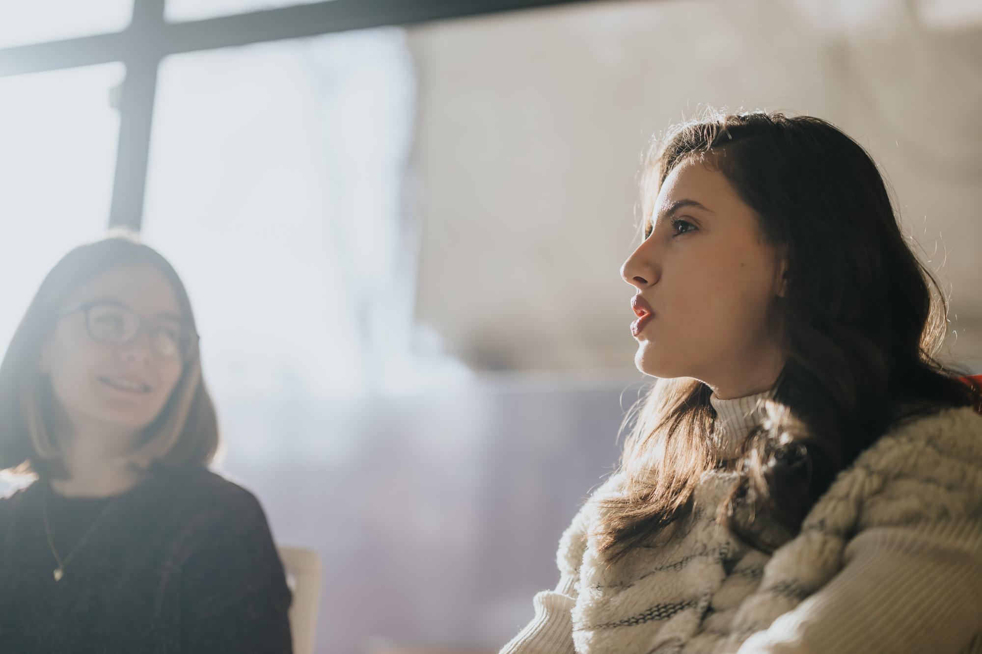 Two women are sitting and talking in a softly lit room. They are engaged in conversation, with one woman in focus as she speaks, while the other listens attentively. Natural light filters in from a large window behind them.