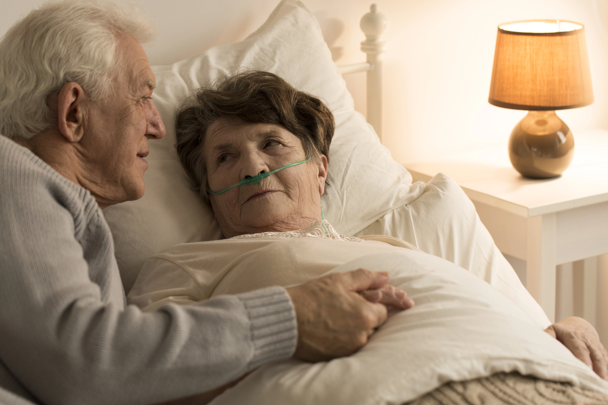 An elderly man sits beside a woman in bed, gently holding her hand. The woman is wearing an oxygen tube. A lamp on a bedside table softly illuminates the room, creating a warm and comforting atmosphere.