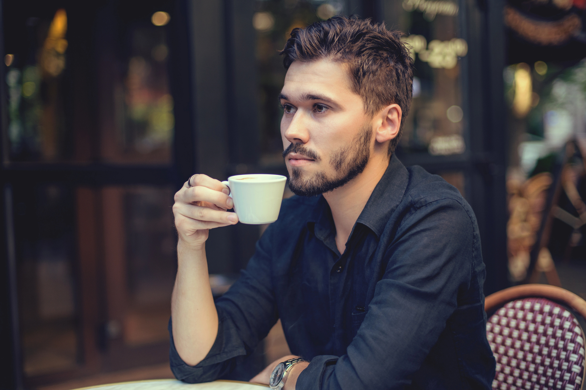 A person with a beard and short hair, wearing a dark shirt, sits at an outdoor cafe table holding a small cup. The backdrop includes a window with blurred bokeh lights. The person looks contemplative, gazing into the distance.