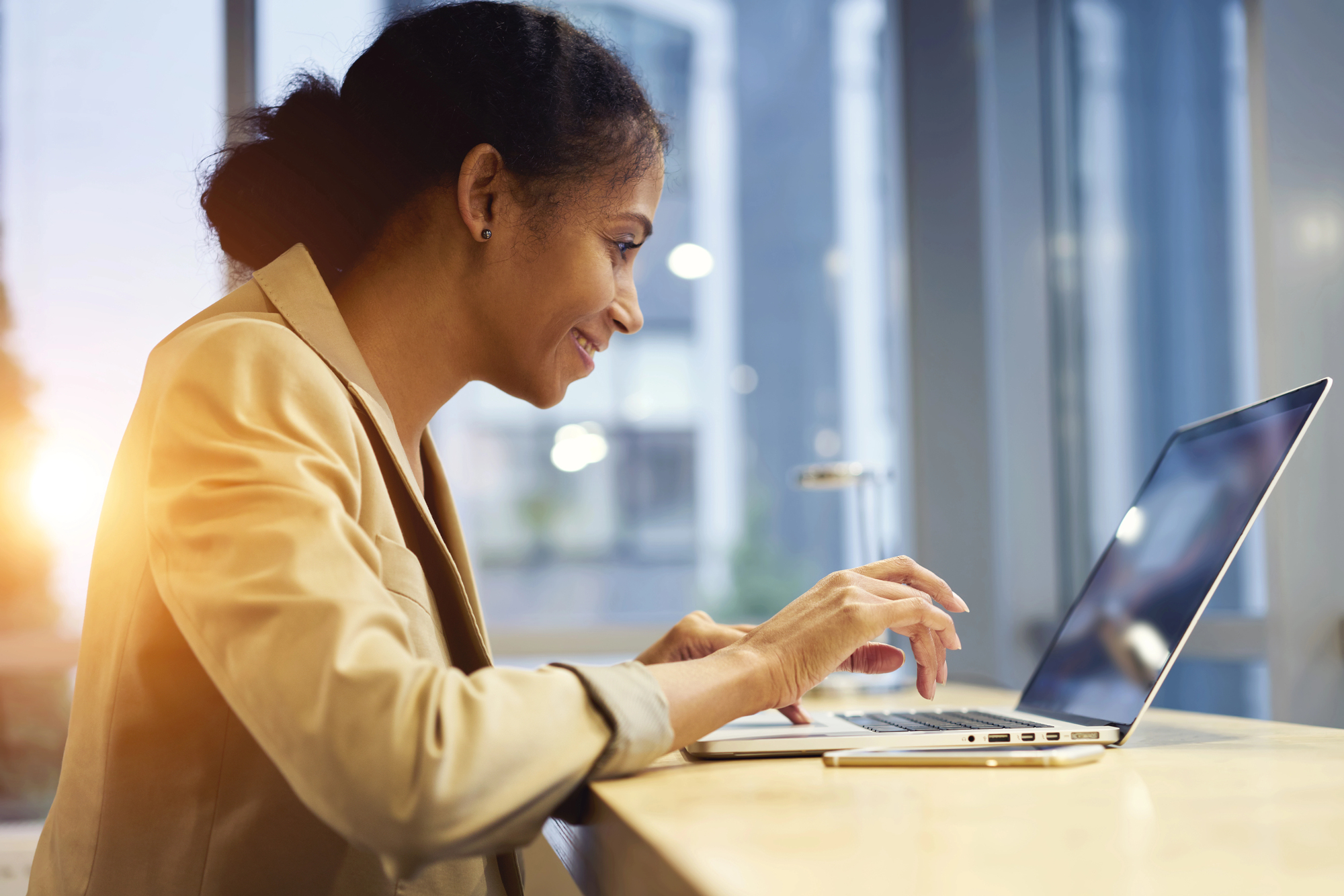 A woman in a tan blazer is sitting at a table, working on a laptop. She is smiling and appears focused. The background shows large windows with a view of a building, and sunlight is streaming in from the left.