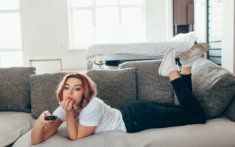 A person with shoulder-length hair, wearing a white t-shirt and jeans, lies on a gray sofa holding a TV remote. They appear relaxed, resting their chin on their hand and legs raised, with a window in the background.