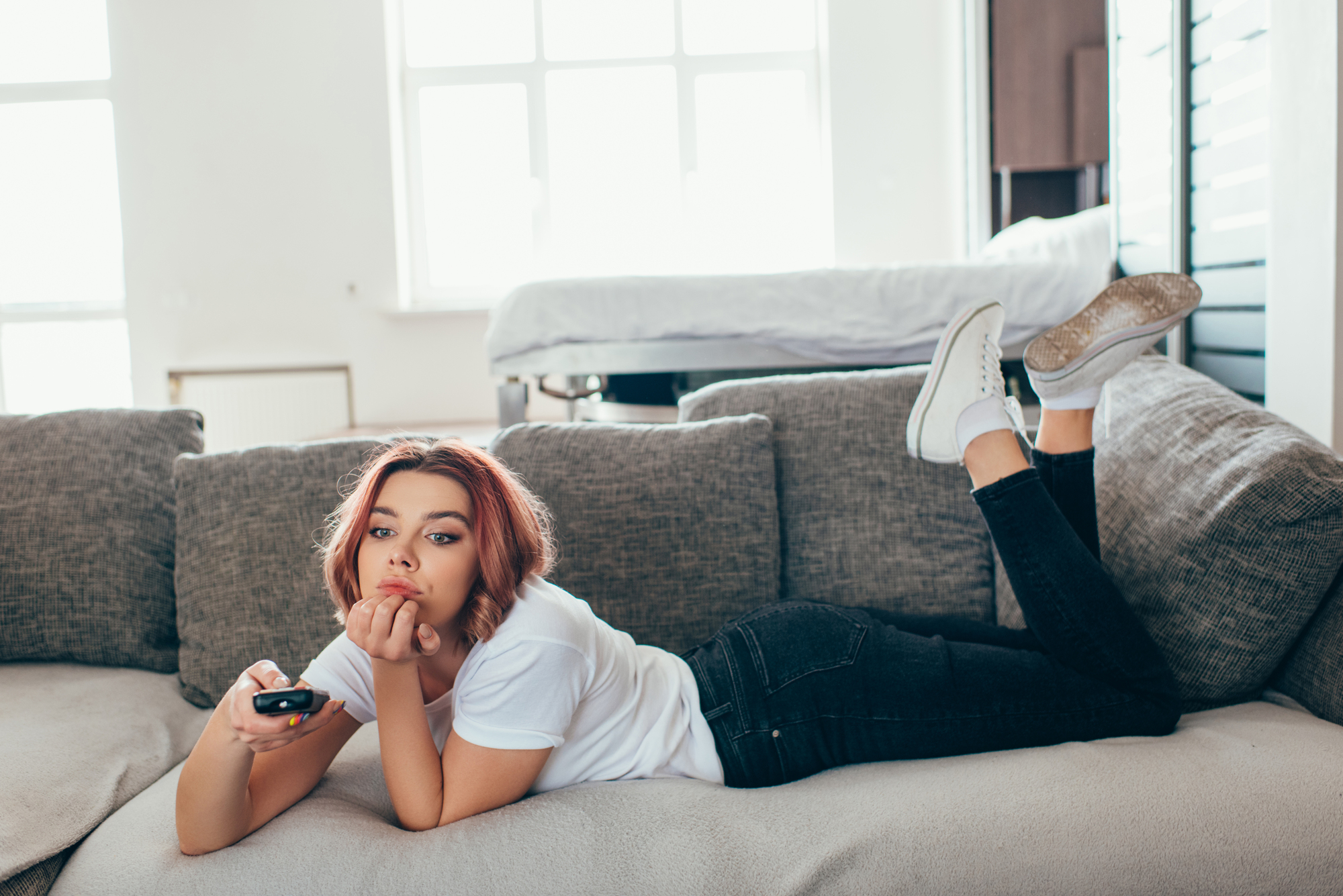 A person with shoulder-length hair, wearing a white t-shirt and jeans, lies on a gray sofa holding a TV remote. They appear relaxed, resting their chin on their hand and legs raised, with a window in the background.