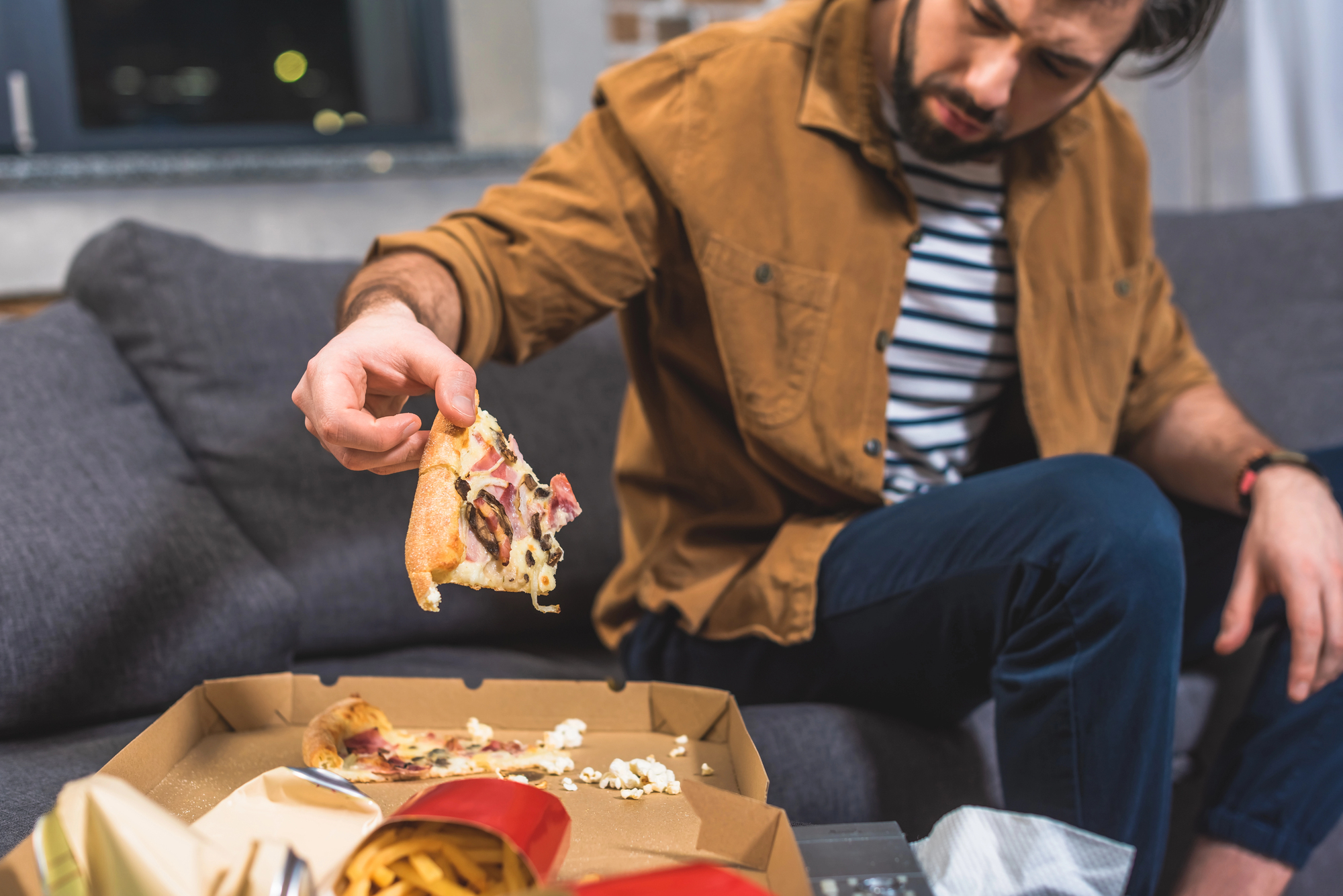 A man in a brown jacket and striped shirt sits on a couch holding a slice of pizza above an open pizza box. There are scattered fries in a container on the table in front of him.