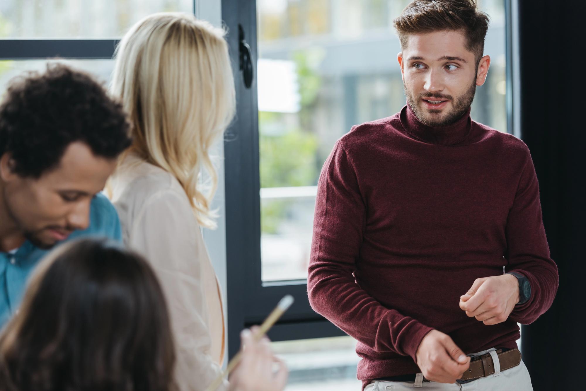 A man in a maroon sweater speaks while gesturing, standing near a seated group in an office setting. A woman with blonde hair and a man with dark curly hair listen. Another person in the foreground holds a pencil. Large windows are in the background.