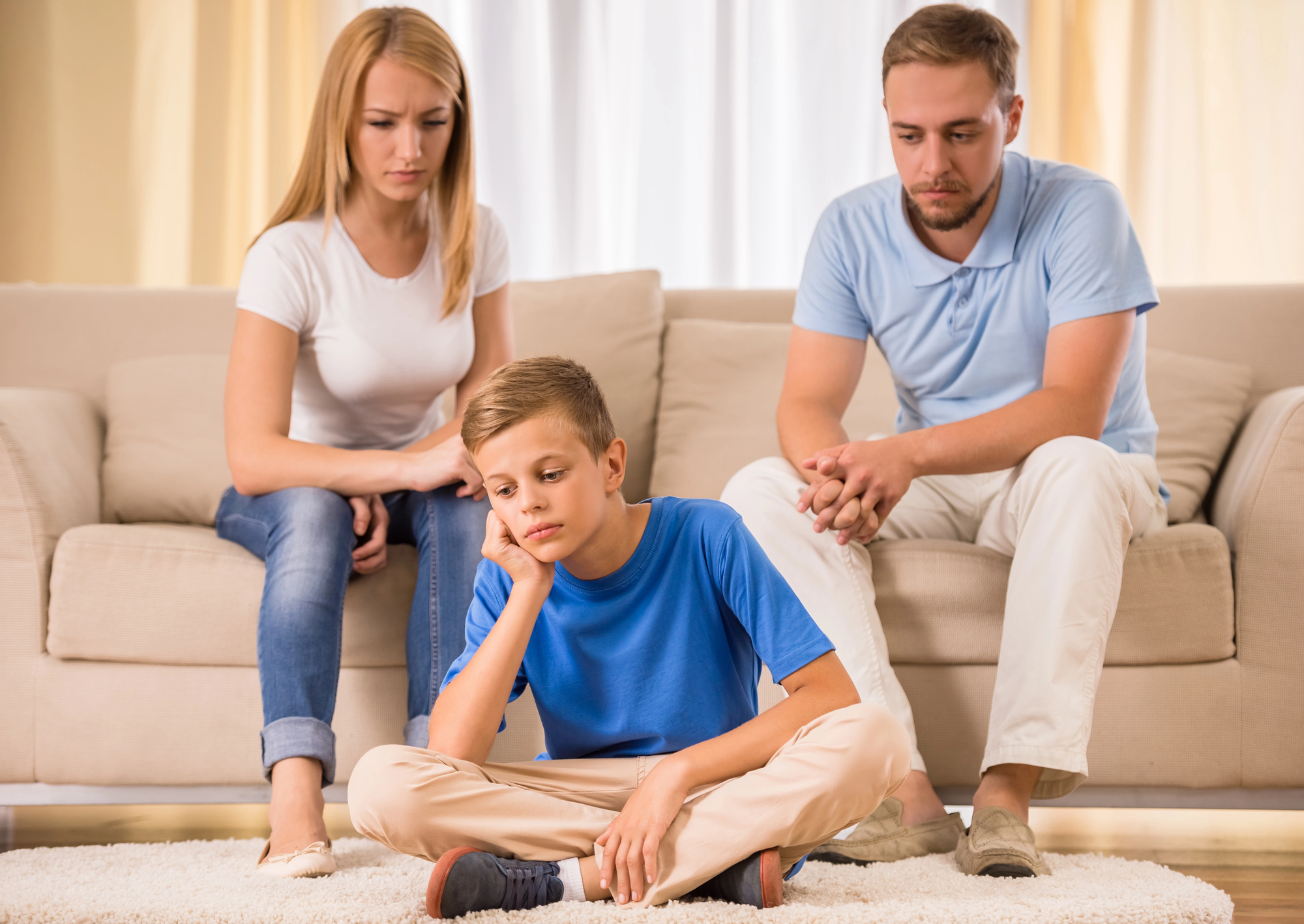 A young boy sits on the floor looking thoughtful, with his chin resting on his hand. Two adults, a woman and a man, sit on a couch behind him, looking concerned. The room is well-lit, with a soothing, neutral color scheme.