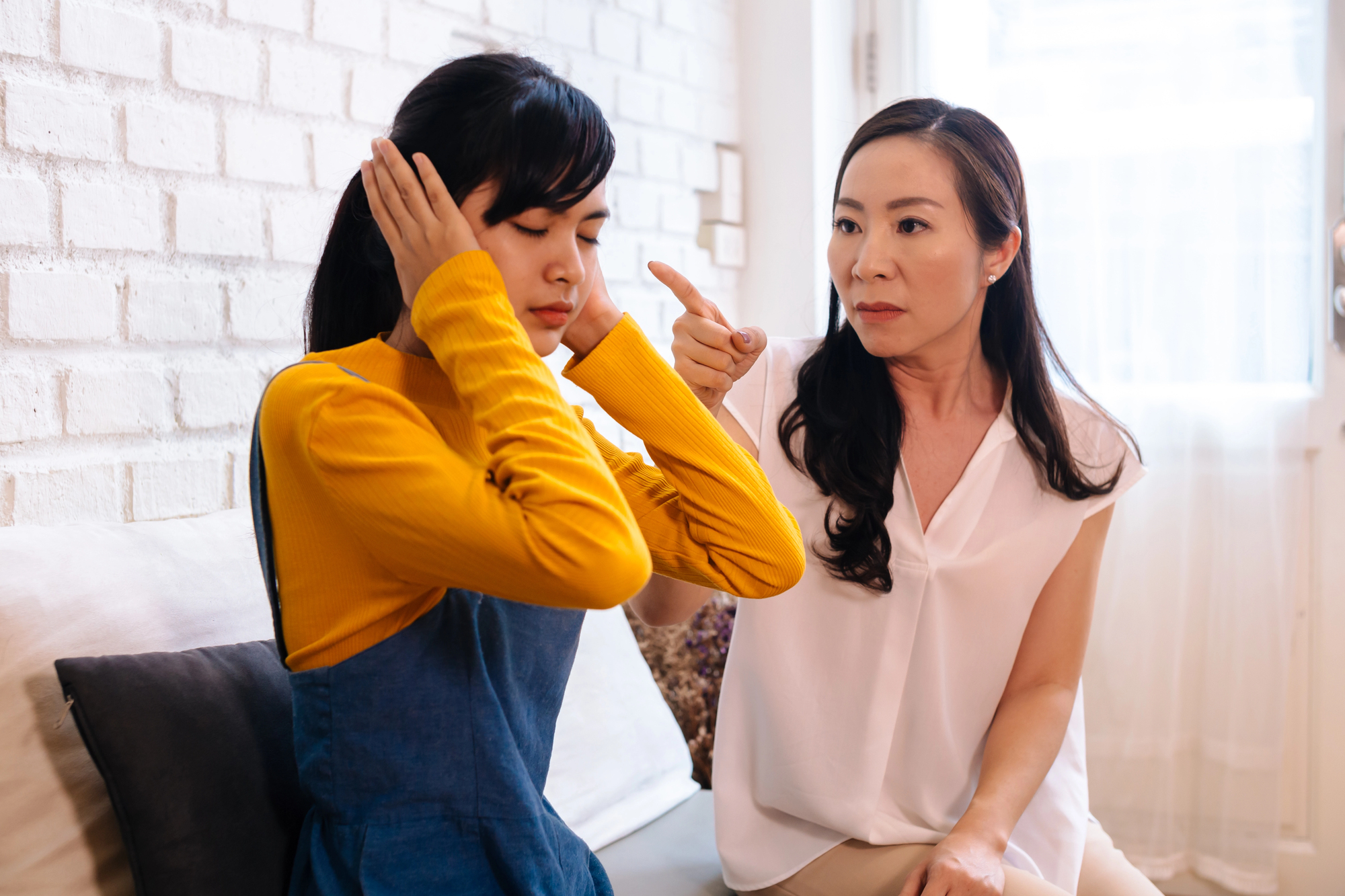 A woman sitting on a couch with eyes closed and hands over ears, while another woman next to her gestures with a pointed finger, appearing frustrated. The scene suggests a disagreement or conflict between them.