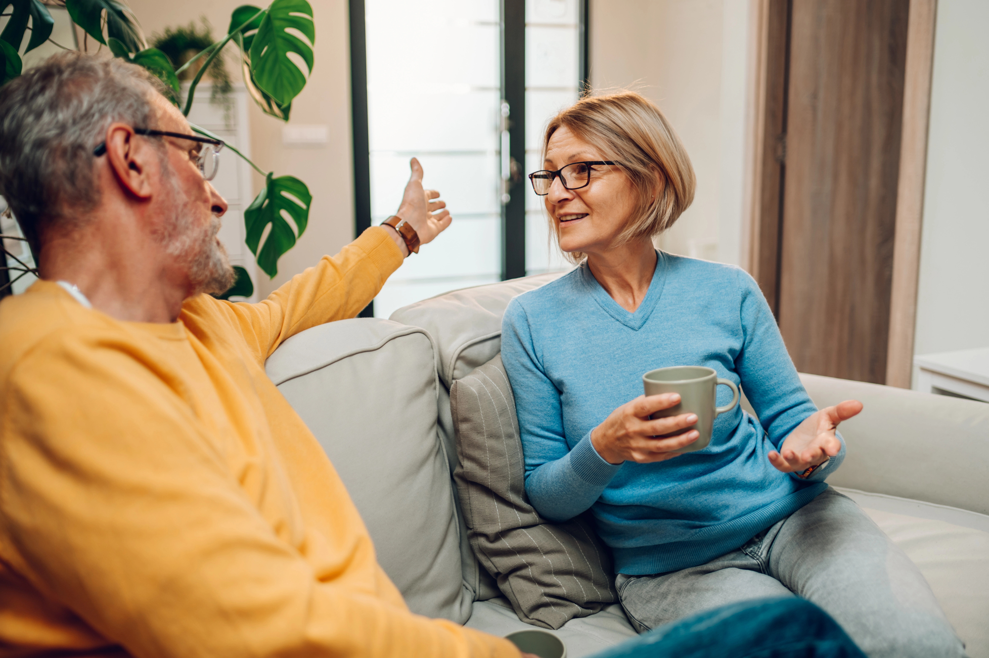 Two people sit on a sofa, engaged in conversation. The man with gray hair wears a yellow sweater, gesturing with his arm. The woman with glasses and a short blonde bob wears a blue sweater, holding a mug. A large plant is in the background.