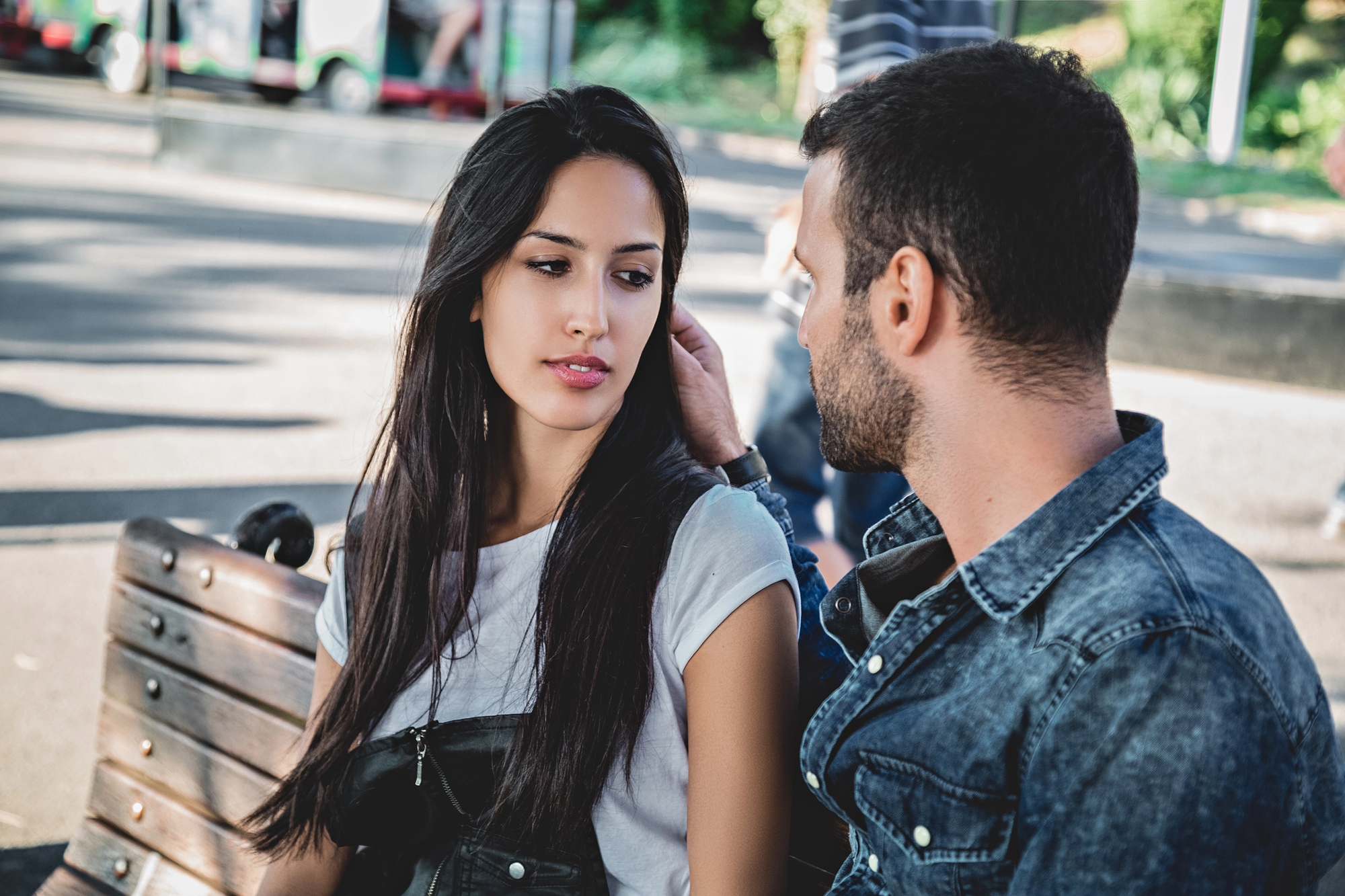 A man and woman sit closely on a park bench. The man, wearing a denim jacket, gently touches the woman's hair. The woman, wearing a short-sleeved top, looks at him with a thoughtful expression. The setting is a sunny outdoor area.