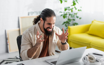 A man with a beard, sitting at a desk, looks frustrated while using a laptop. He raises his hands in exasperation. There is a bright yellow sofa and a potted plant in the background. Headphones and a smartphone are on the desk.