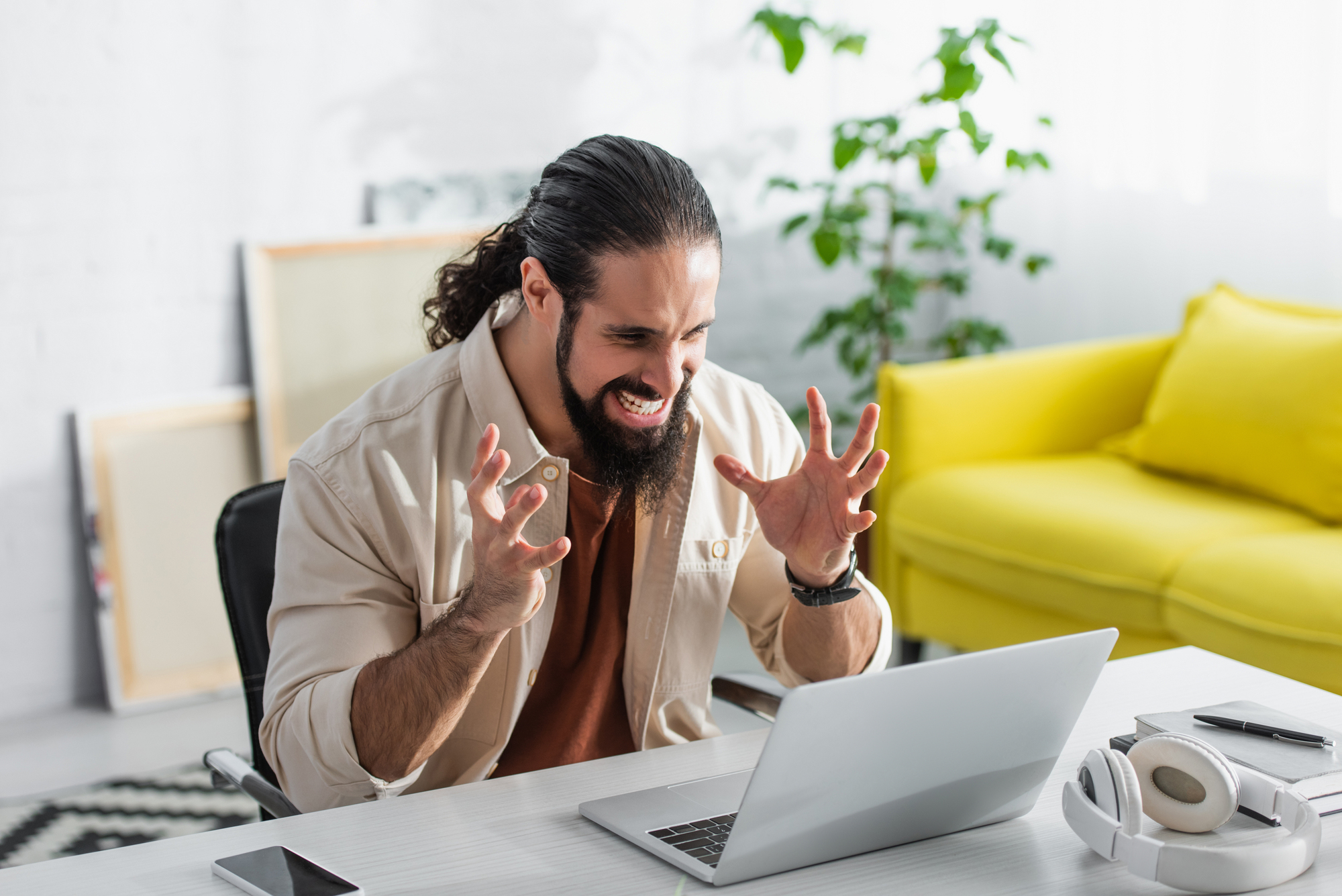 A man with a beard, sitting at a desk, looks frustrated while using a laptop. He raises his hands in exasperation. There is a bright yellow sofa and a potted plant in the background. Headphones and a smartphone are on the desk.