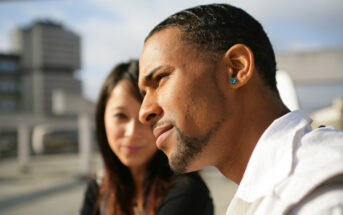 A man and a woman are sitting outdoors in a city setting under a clear sky. The man in the foreground is wearing a white shirt and has an earring, while the woman in the background is smiling slightly. Buildings are visible behind them.