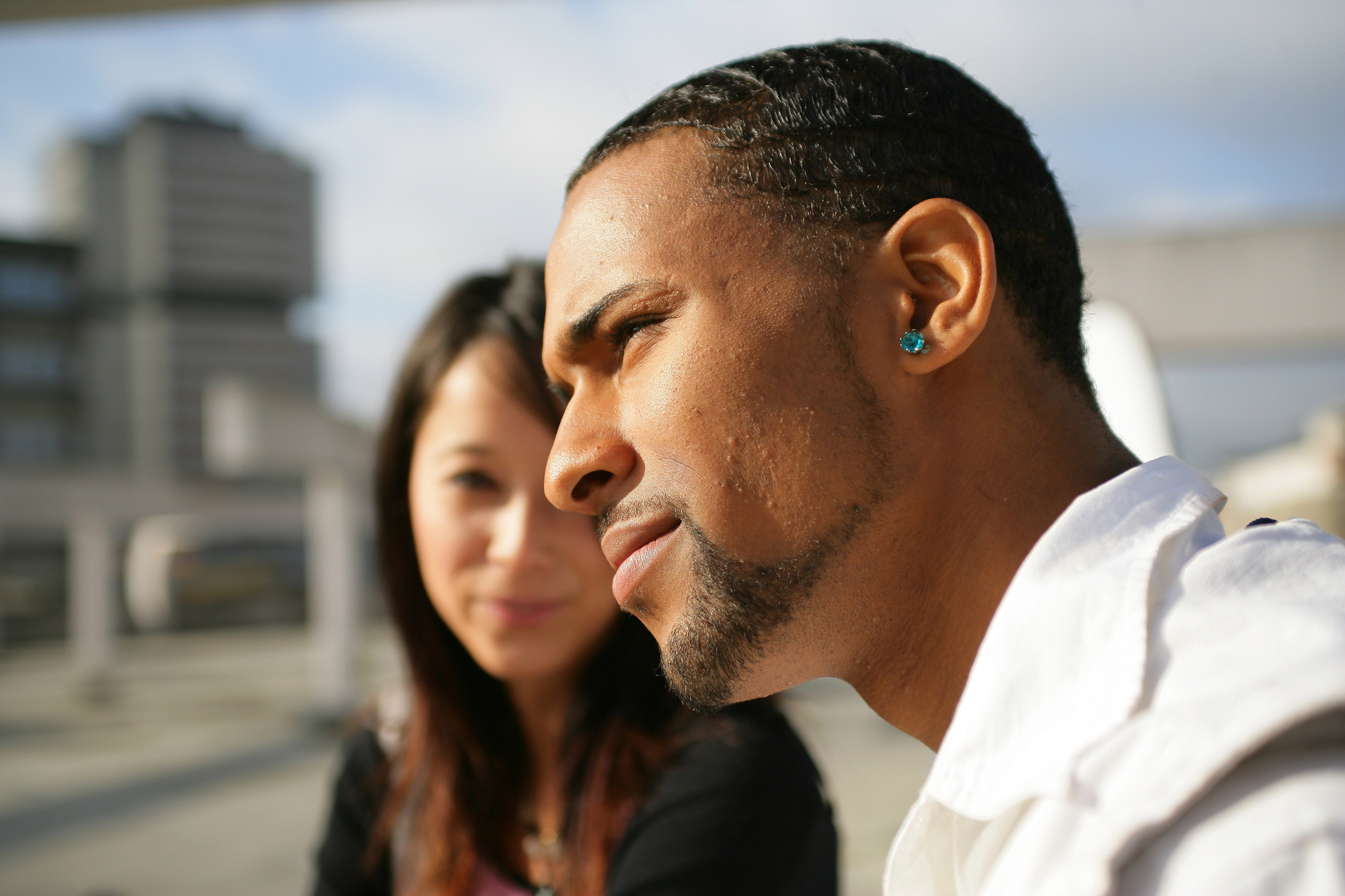 A man and a woman are sitting outdoors in a city setting under a clear sky. The man in the foreground is wearing a white shirt and has an earring, while the woman in the background is smiling slightly. Buildings are visible behind them.