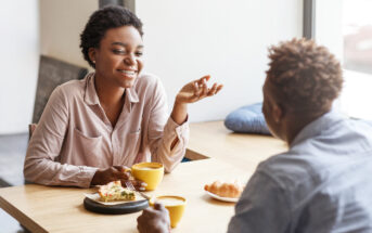 Two people sit at a wooden table by a window, enjoying coffee and pastries. The woman is smiling, gesturing with her hand while holding a coffee cup. The man faces her, listening attentively. Sunlight filters through the window, creating a warm ambiance.