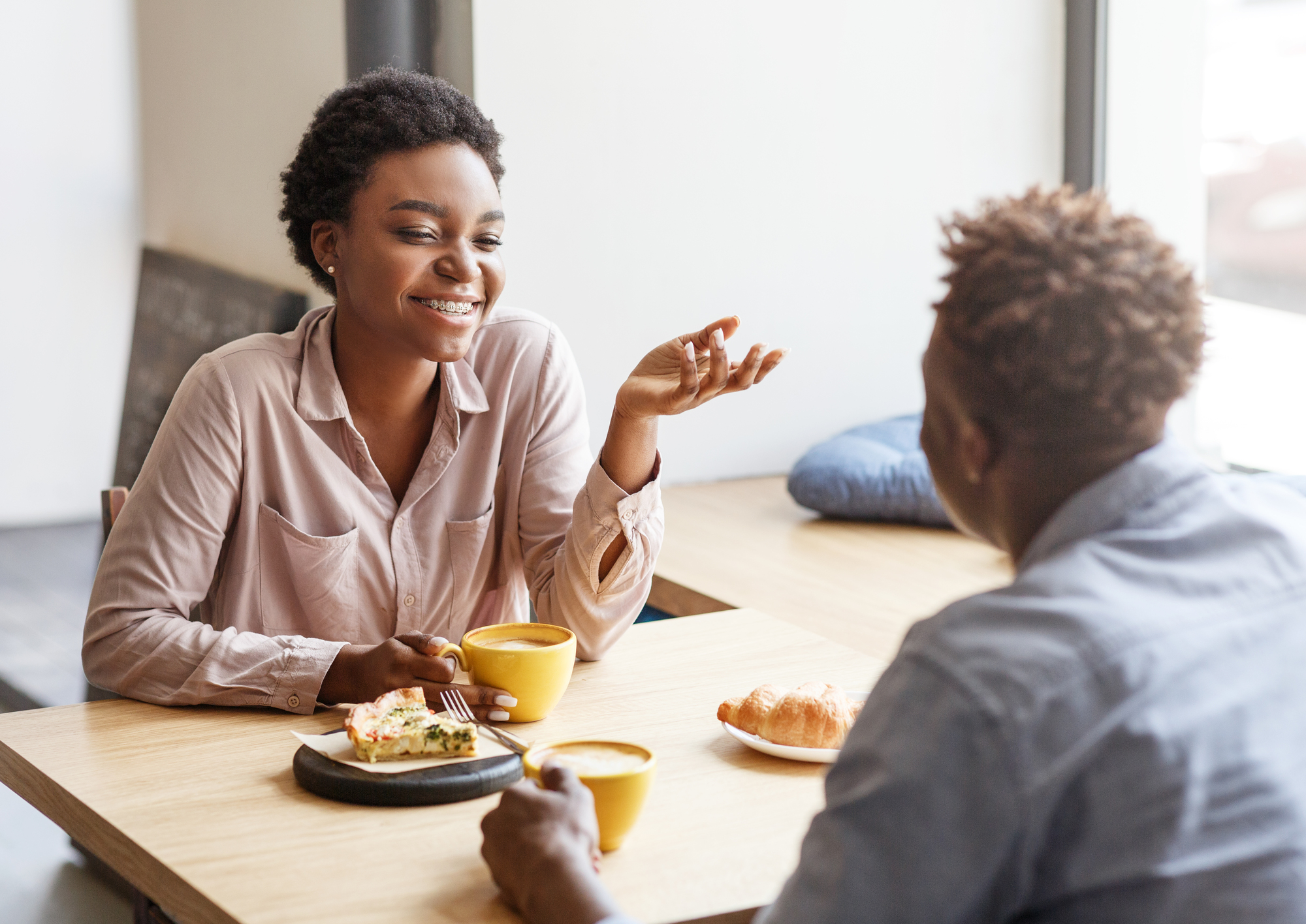 Two people sit at a wooden table by a window, enjoying coffee and pastries. The woman is smiling, gesturing with her hand while holding a coffee cup. The man faces her, listening attentively. Sunlight filters through the window, creating a warm ambiance.