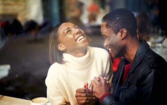 A couple sitting at a cafe table, holding hands and laughing joyfully. The woman is wearing a white sweater, and the man is in a black jacket. A cup of coffee is on the table. The background is softly blurred, suggesting a cozy, bustling atmosphere.