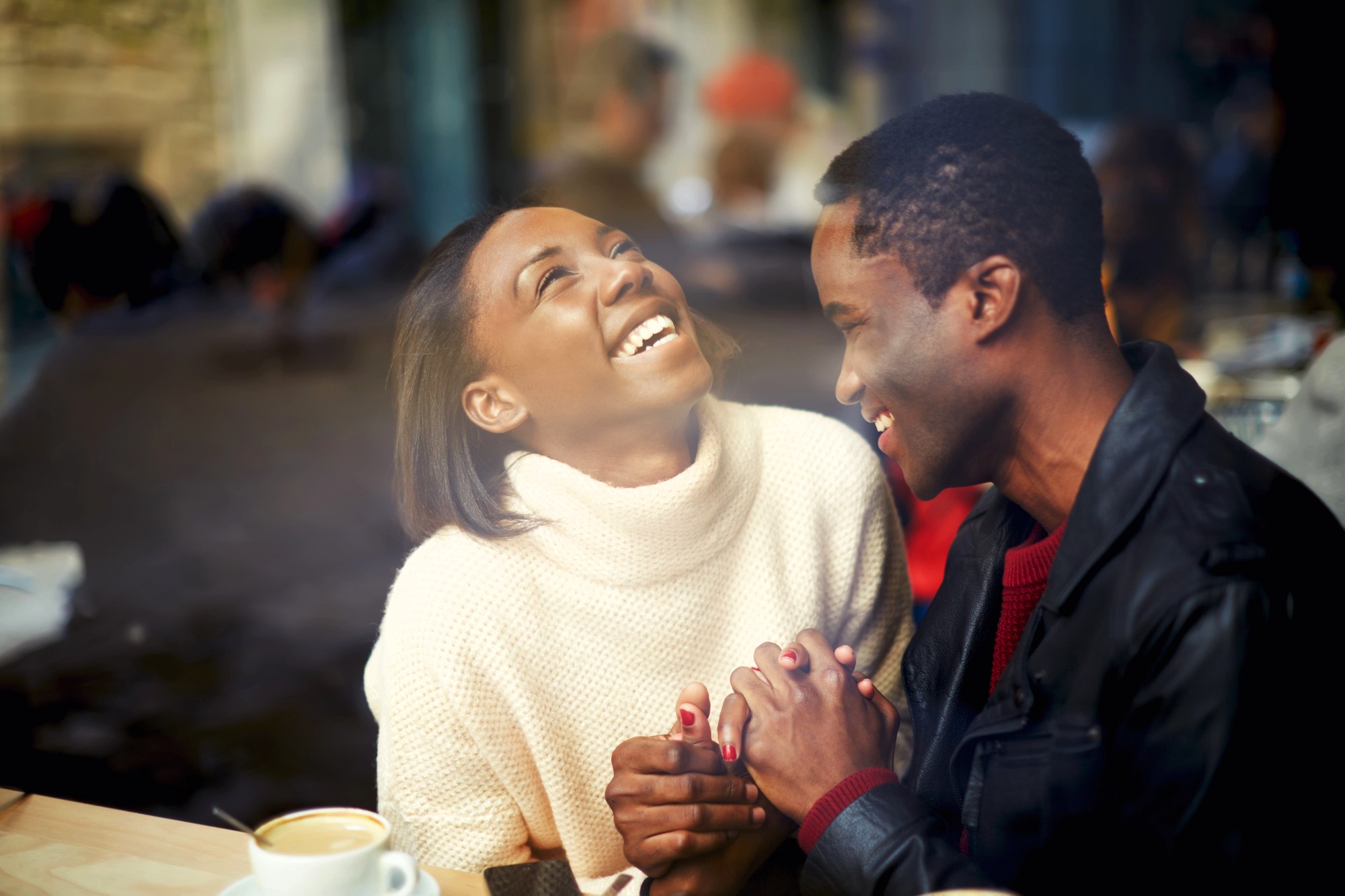 A couple sitting at a cafe table, holding hands and laughing joyfully. The woman is wearing a white sweater, and the man is in a black jacket. A cup of coffee is on the table. The background is softly blurred, suggesting a cozy, bustling atmosphere.