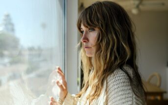 A woman with long hair and a knitted sweater gazes thoughtfully out a window. Her hands lightly touch the glass. The room is softly lit, revealing a chair and table in the background. The scene conveys a contemplative mood.