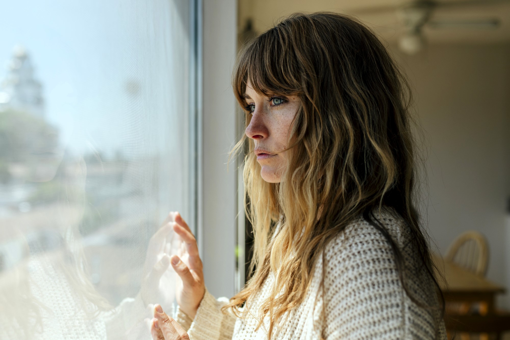 A woman with long hair and a knitted sweater gazes thoughtfully out a window. Her hands lightly touch the glass. The room is softly lit, revealing a chair and table in the background. The scene conveys a contemplative mood.