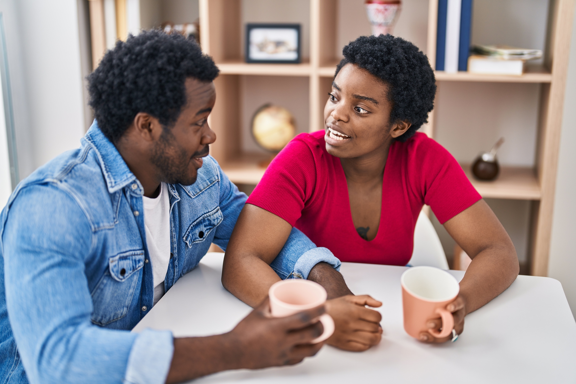 A man and a woman sit at a table holding mugs and smiling at each other. The man wears a denim shirt, and the woman wears a red top. Books and decorative items are visible on a shelf in the background.